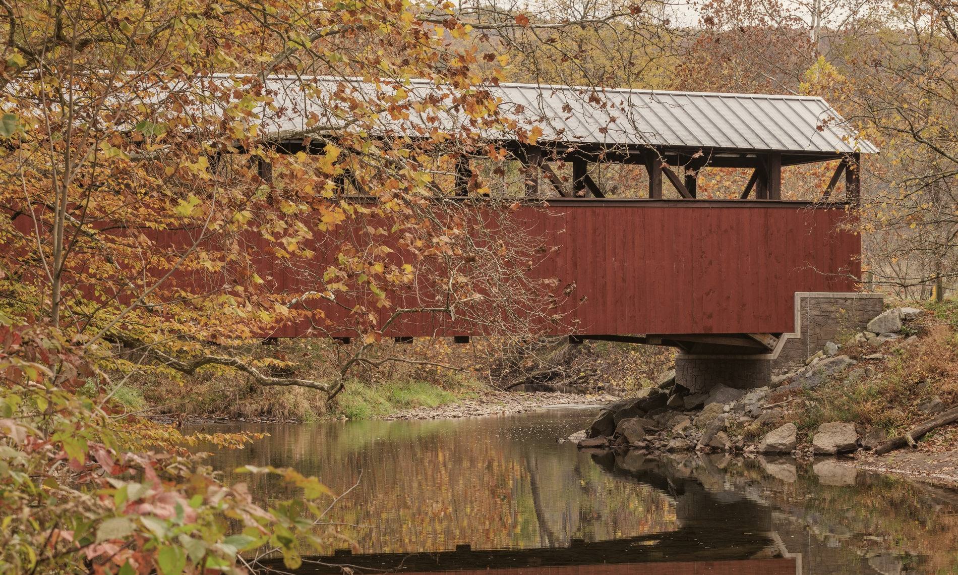 A red covered bridge over a still river among autumn trees, mirrored in the water.