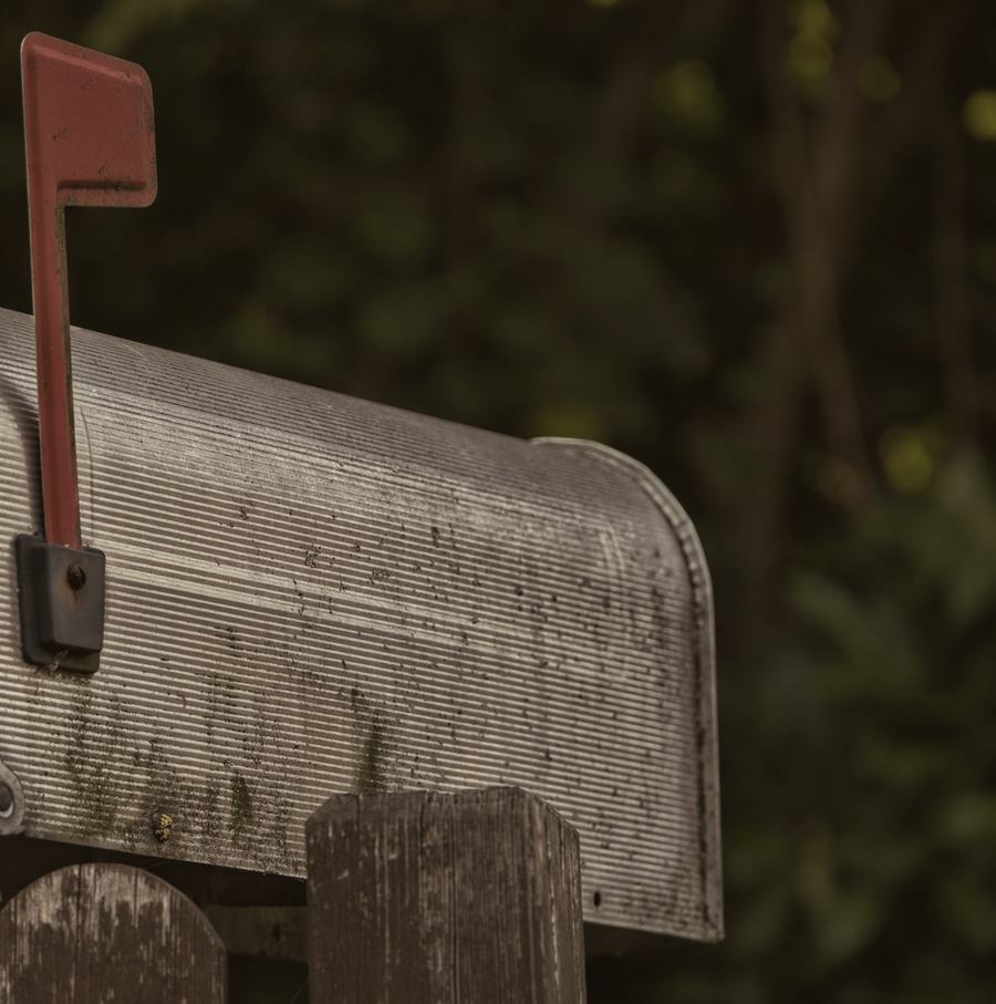 The side of a weathered metal rural mailbox on a wooden post, its red flag raised.