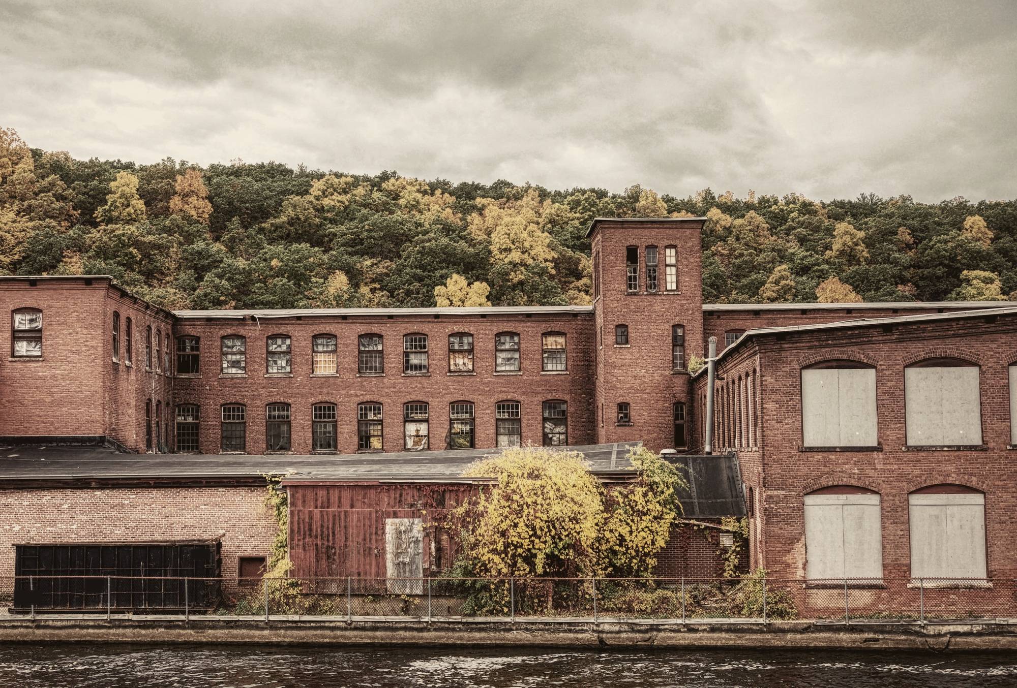 Brick mill buildings with rows of arched windows and a stair tower on a riverbank, a wooded hill behind with a few trees turning yellow.